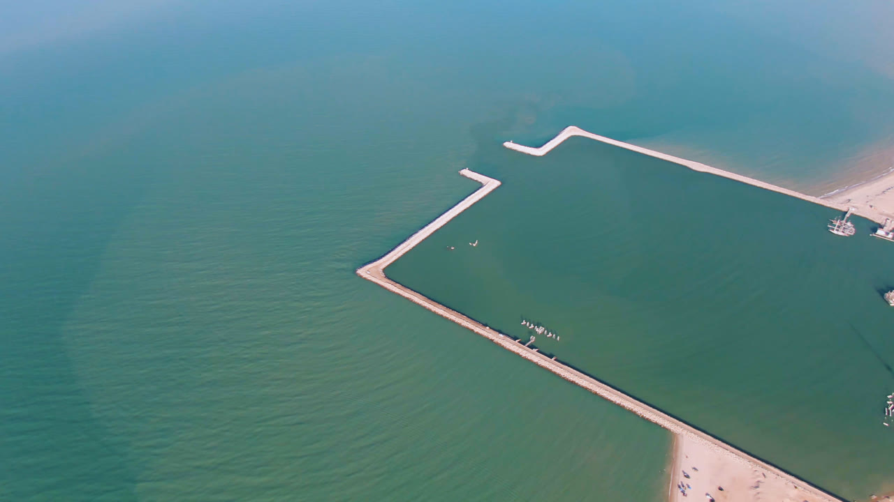 Opening aerial frame over San Felipe, Mexico, showing a pale angular pier across green-blue water with sandy beach at the lower right and no visible people.