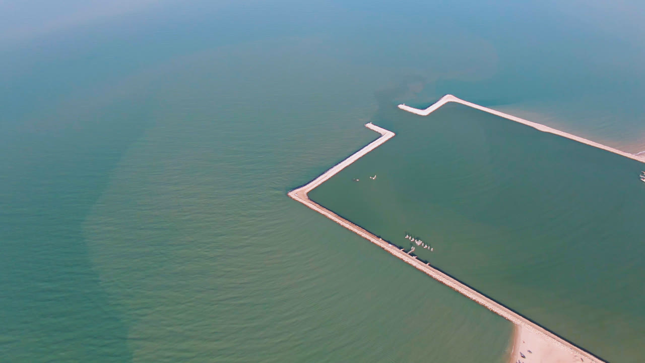 High aerial frame of a pale pier crossing green water with beach at the lower edge.