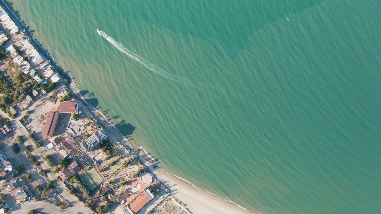 Overhead coastline with turquoise water, sandy beach, buildings, and a boat wake.