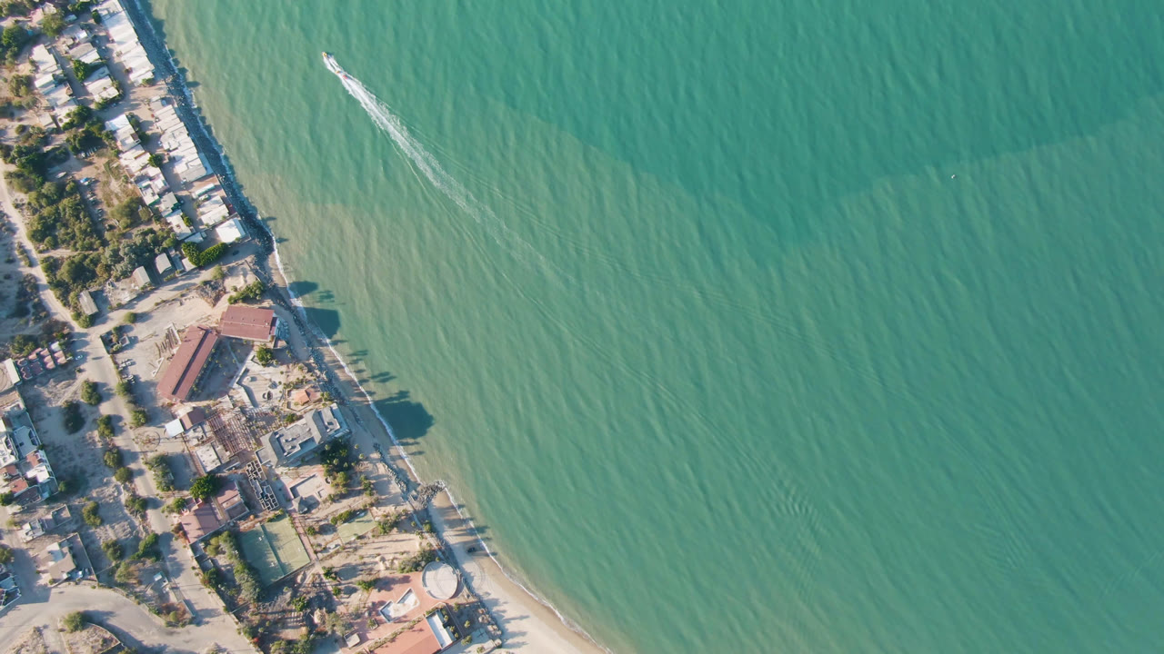 High aerial view of San Felipe, Mexico coastline with green water filling the right side, beach and low buildings along the left, a small boat wake near shore, and no visible people.