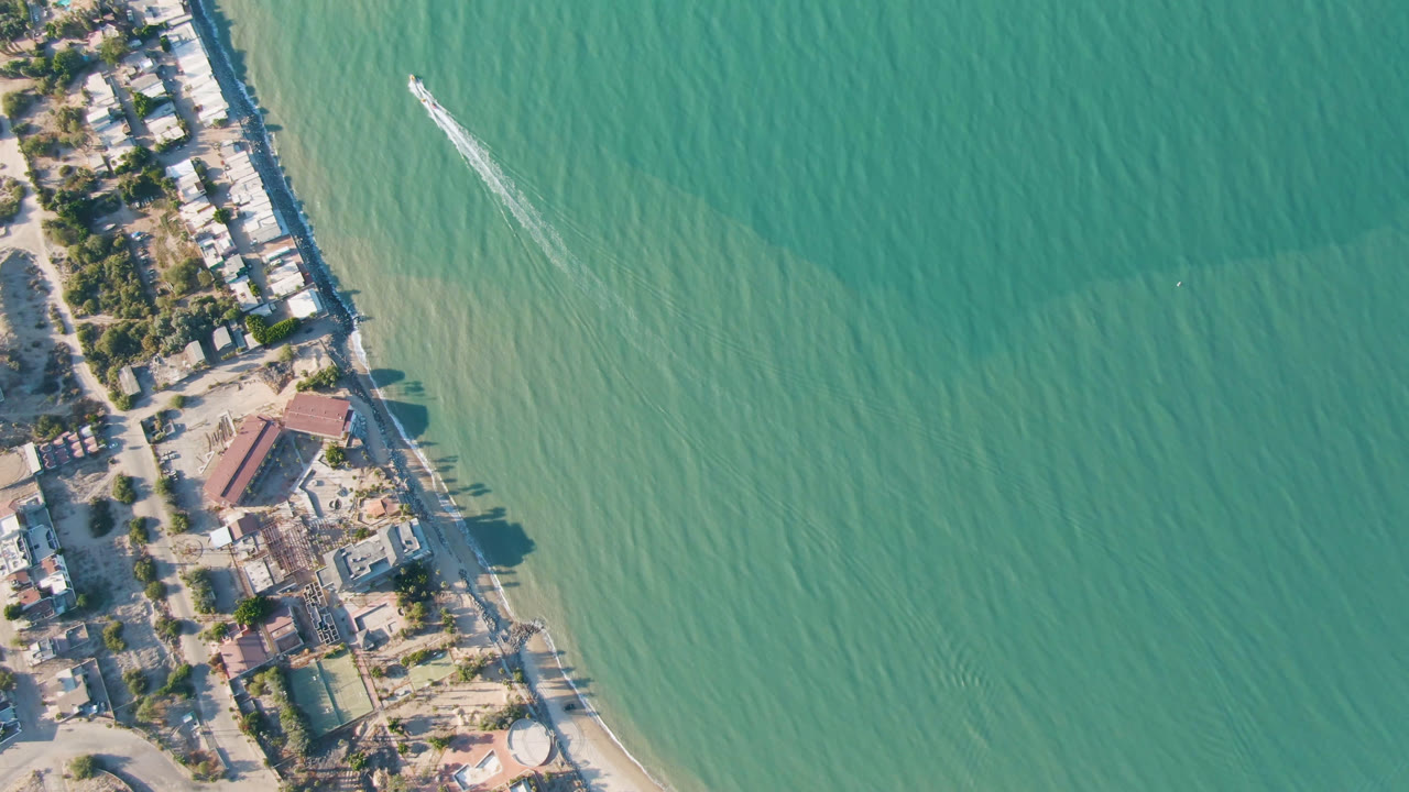 Drone view of diagonal coast with beach buildings, trees, open water, and a boat wake.