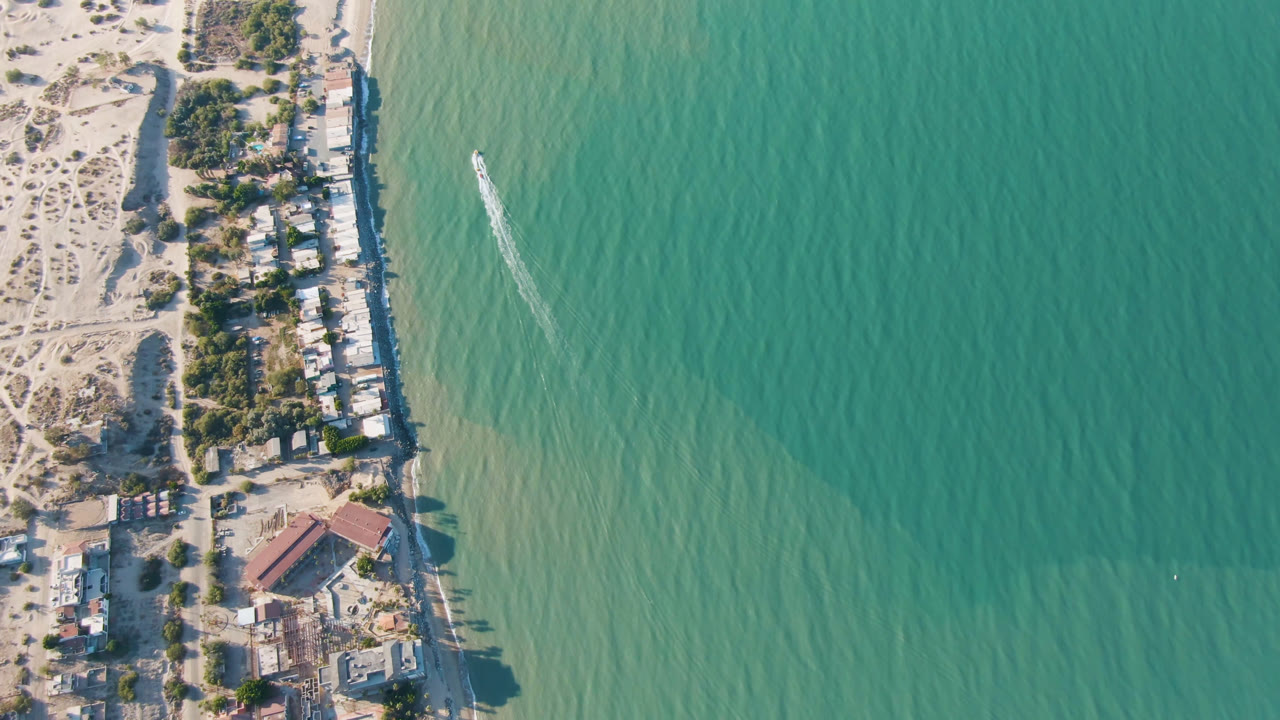 Overhead beach edge with shoreline buildings, trees, turquoise water, and a small boat wake.