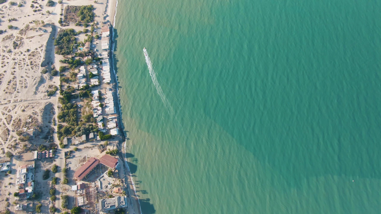 Top-down San Felipe coastal frame with a row of beachside buildings and trees at left, shallow green water at center, and a small boat track near the shore with no visible people.