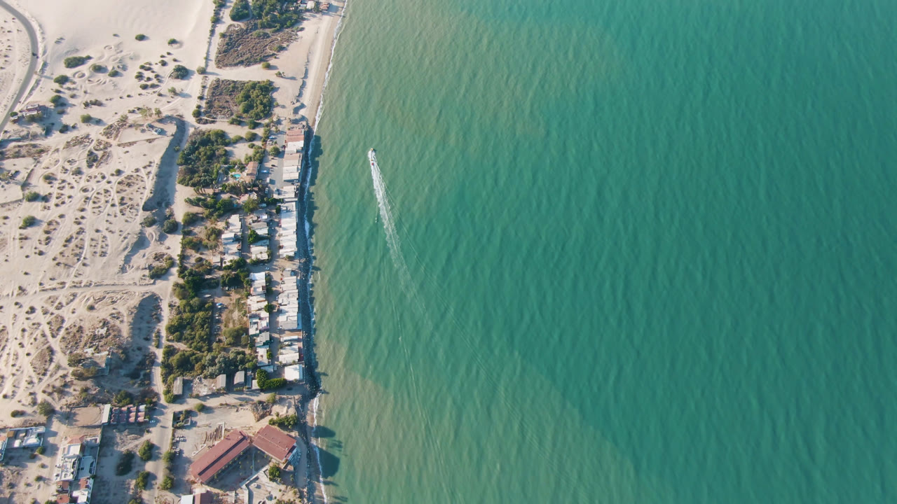 Aerial frame over San Felipe, Baja California, Mexico, with pale dunes, a line of shoreline houses, turquoise water, a small boat wake offshore, and no visible people.