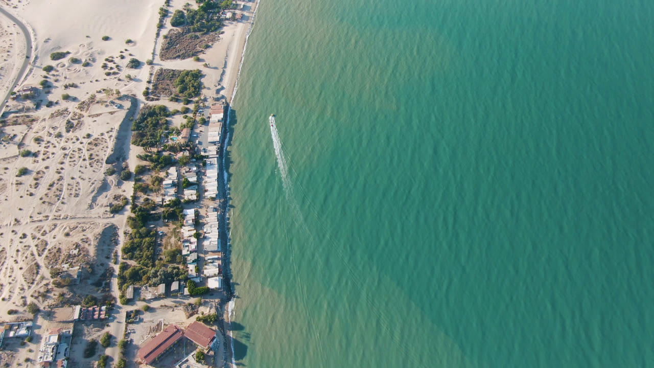 A high drone view of San Felipe coastline shows dune tracks, beach houses and trees along the shore, green-blue water to the right, a narrow white wake, and no visible people.