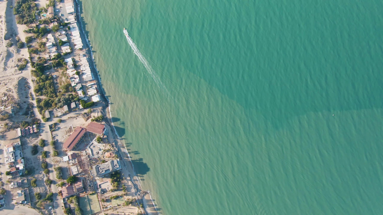 Overhead coastline with turquoise water, beach homes, sand, and a small boat wake.