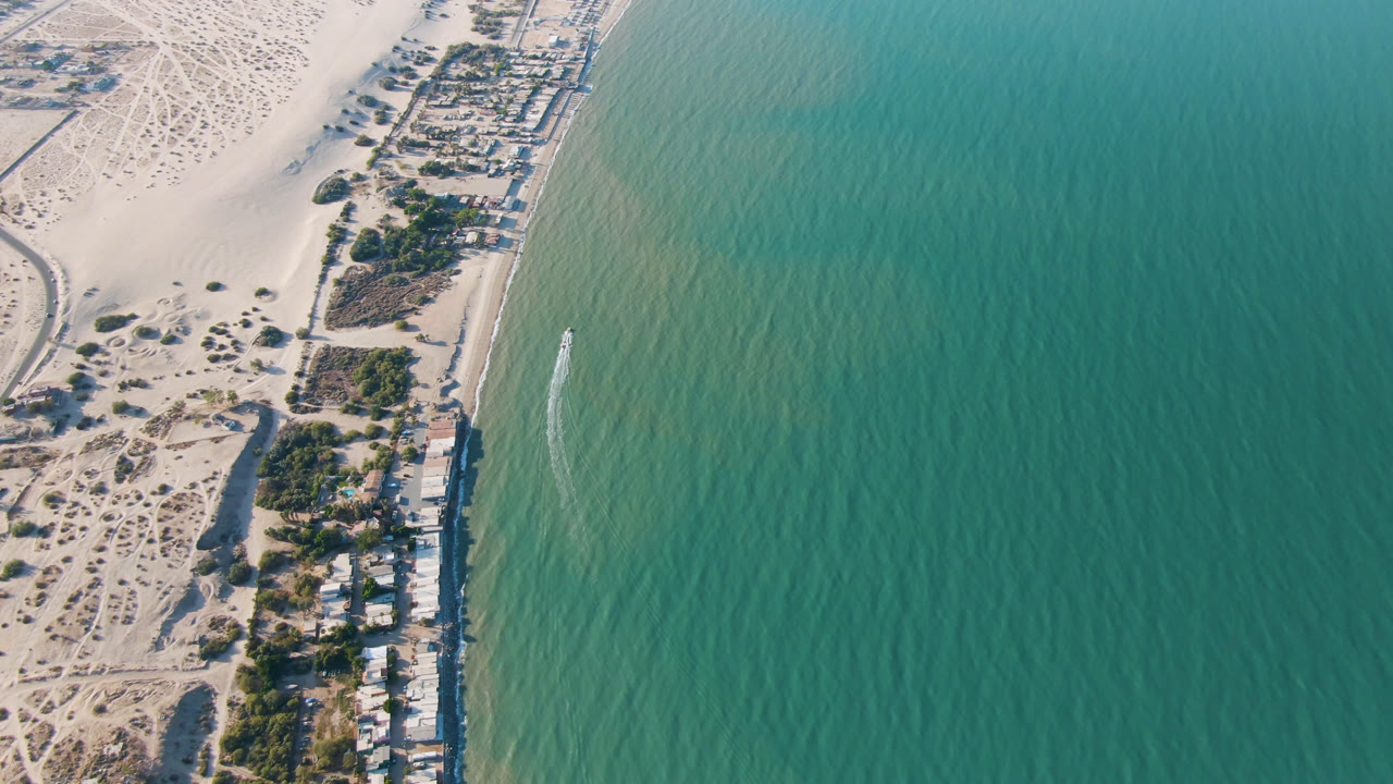 High aerial frame of San Felipe coastline showing beach homes beside pale sand, open green-blue water, a small offshore wake, and no visible people.