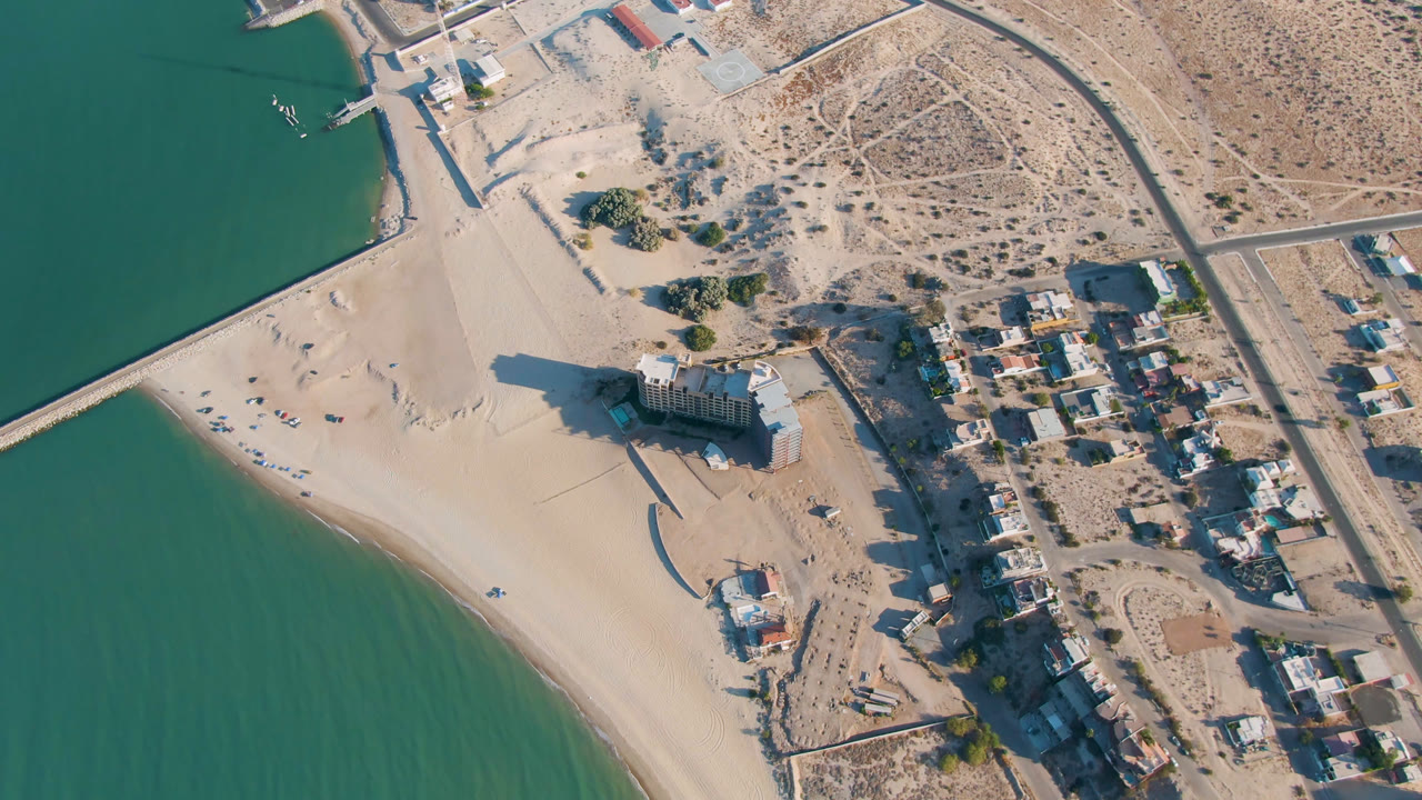 Aerial frame over San Felipe, Baja California showing green coastal water, a wide sandy beach, a towered waterfront building, nearby roads, small structures, desert terrain, and no visible people.