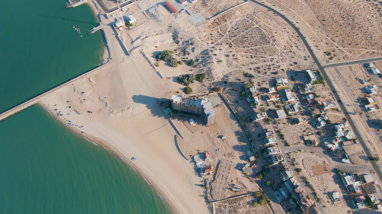 Second aerial frame over San Felipe, Baja California with the beach and green water at left, the towered building near the shoreline, small boats by a pier, inland roads, and no visible people.
