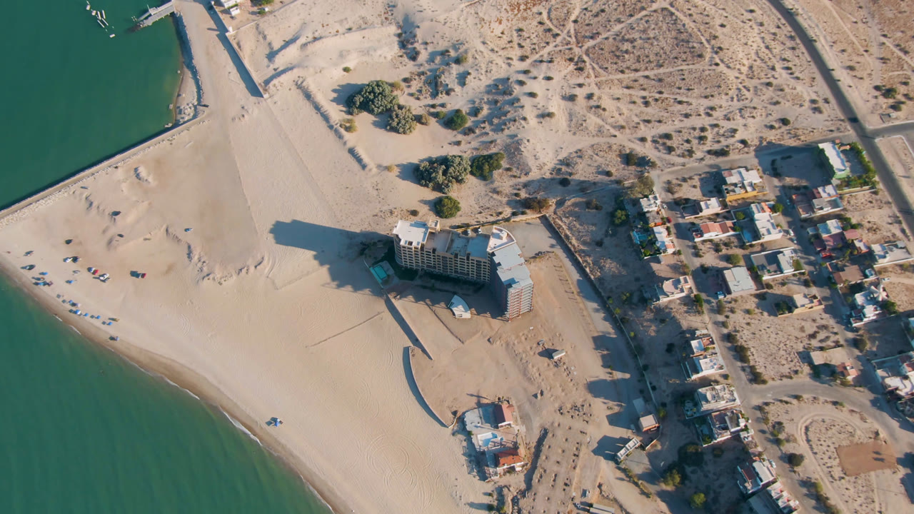 Overhead view of a sandy beach, green water, beachfront hotel, roads, and desert housing.