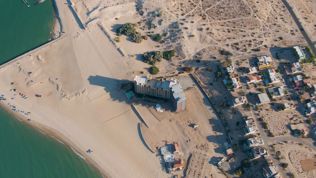 Second aerial frame over San Felipe beach, showing the hotel slightly lower in frame, pale sand around it, green water at left, a road grid and small homes to the right, and no visible people.