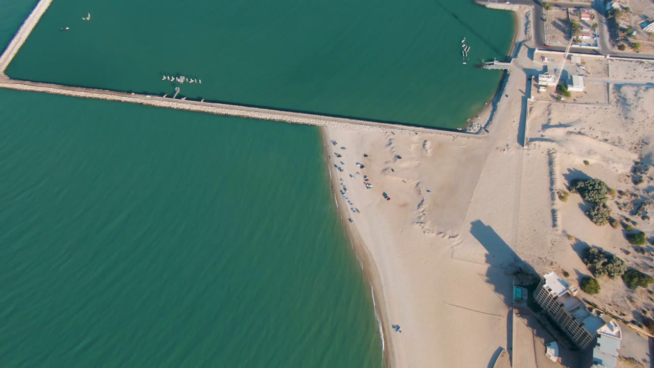 High aerial frame over San Felipe, Baja California showing the sandy beach between green water and harbor walls, with the pier house casting a long shadow and no visible people.