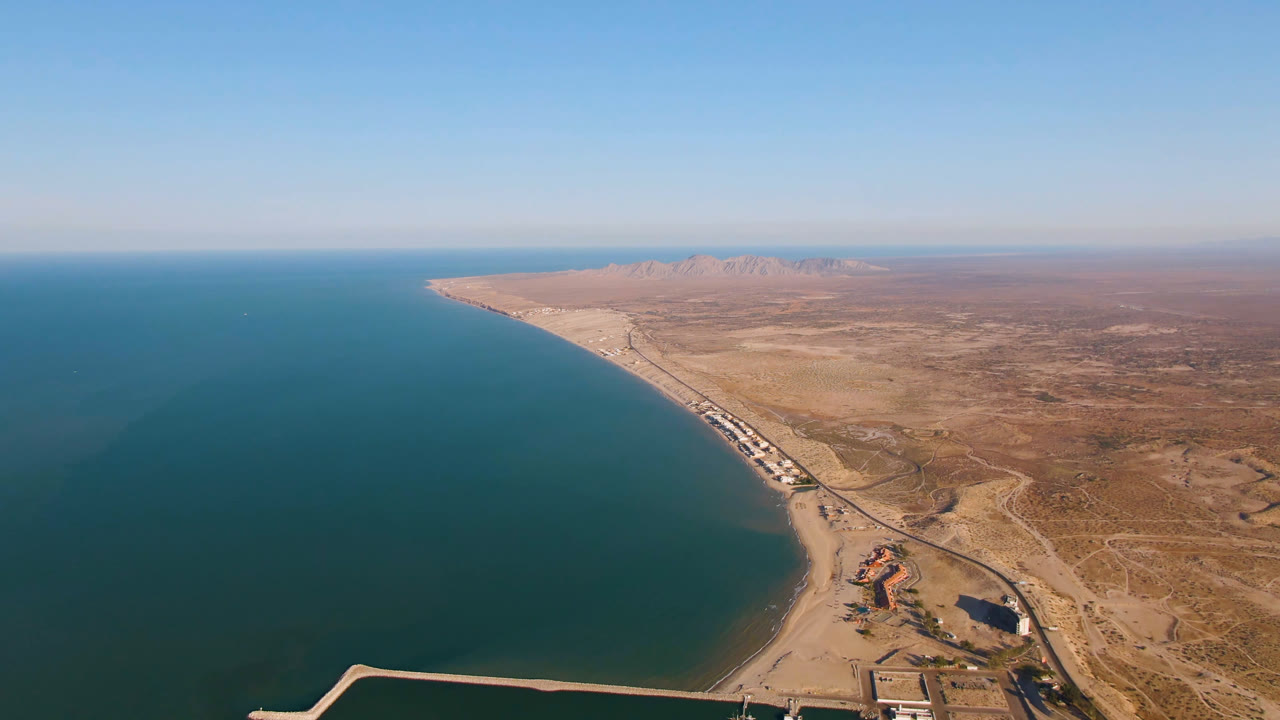 Wide aerial frame of San Felipe, Baja California coastline with blue-green water on the left, tan beach and desert on the right, distant mountains on the horizon, and no visible people.