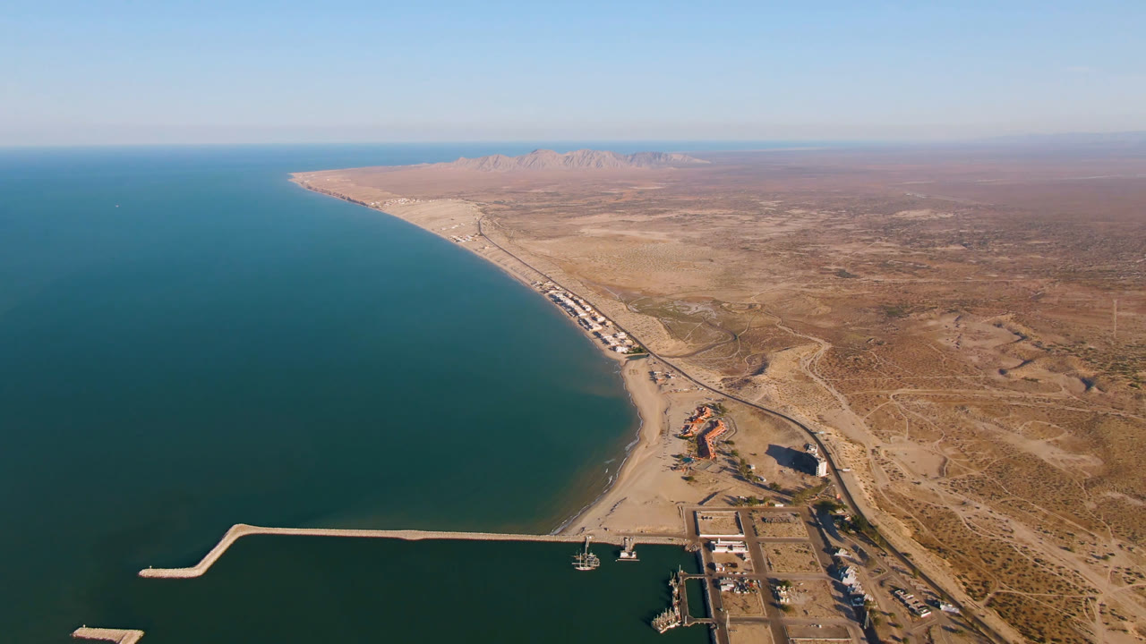 Aerial frame over San Felipe, Mexico showing teal coastal water beside a long sandy beach, a harbor breakwater in the foreground, dry inland terrain, distant mountains, and no visible people.