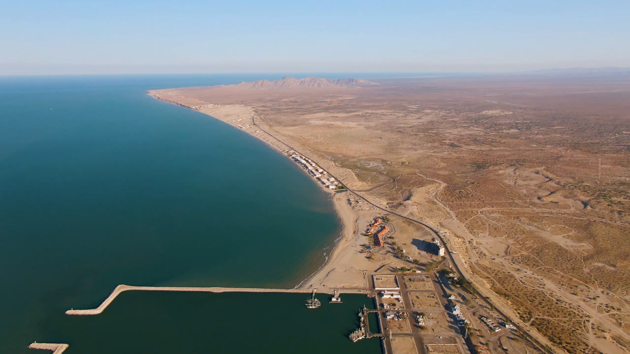 Aerial frame over San Felipe, Mexico with the harbor breakwater lower in frame, teal water along the left side, sandy shoreline running toward distant mountains, and no visible people.