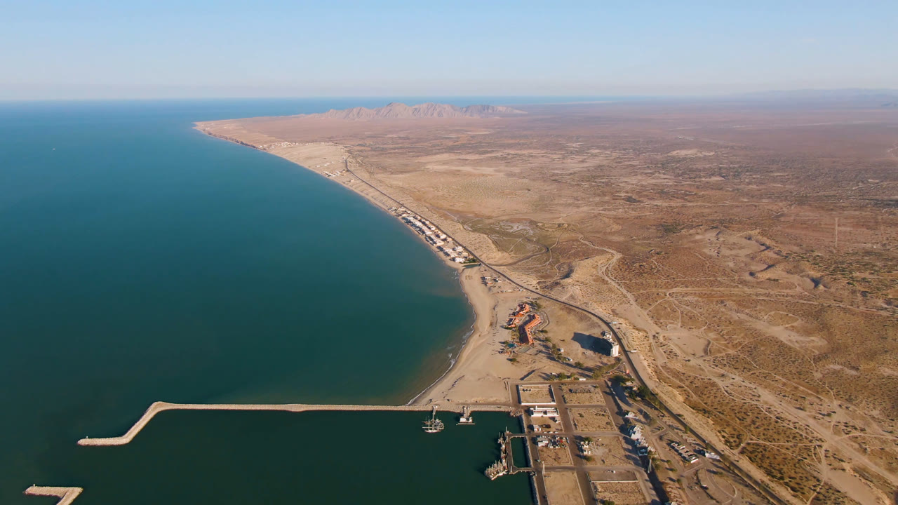 Aerial coastline with teal water, breakwater, beach road, desert, and distant mountains.