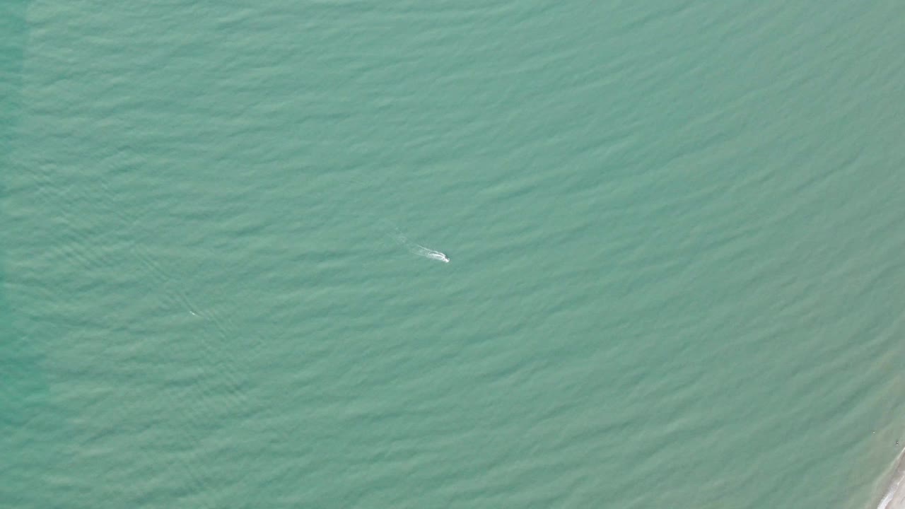 Aerial view over turquoise ocean in San Felipe, BC, Mexico, with a small white kite near the upper left area, rippled water below, a sliver of shoreline on the right edge, and no visible people.