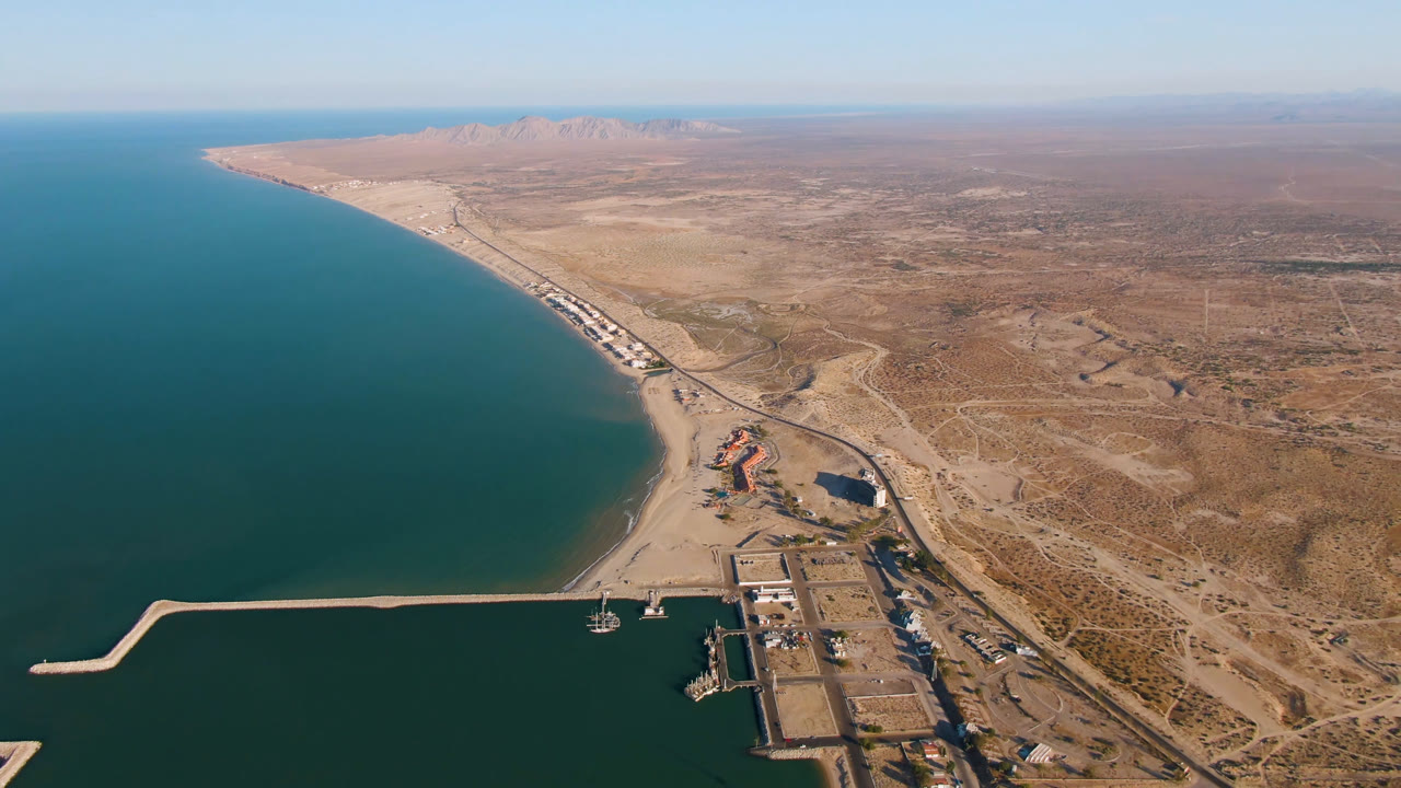 Aerial view over San Felipe, Mexico showing turquoise water, a curved beach, harbor breakwater, dry inland terrain, distant mountains, and no visible people at the start of the clip.