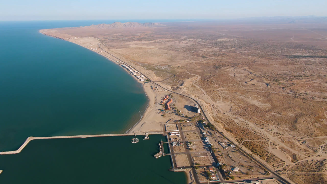 Aerial view of San Felipe, Mexico with deep teal water on the left, the harbor and breakwater below, pale beach beyond, dry desert roads to the right, and no visible people.