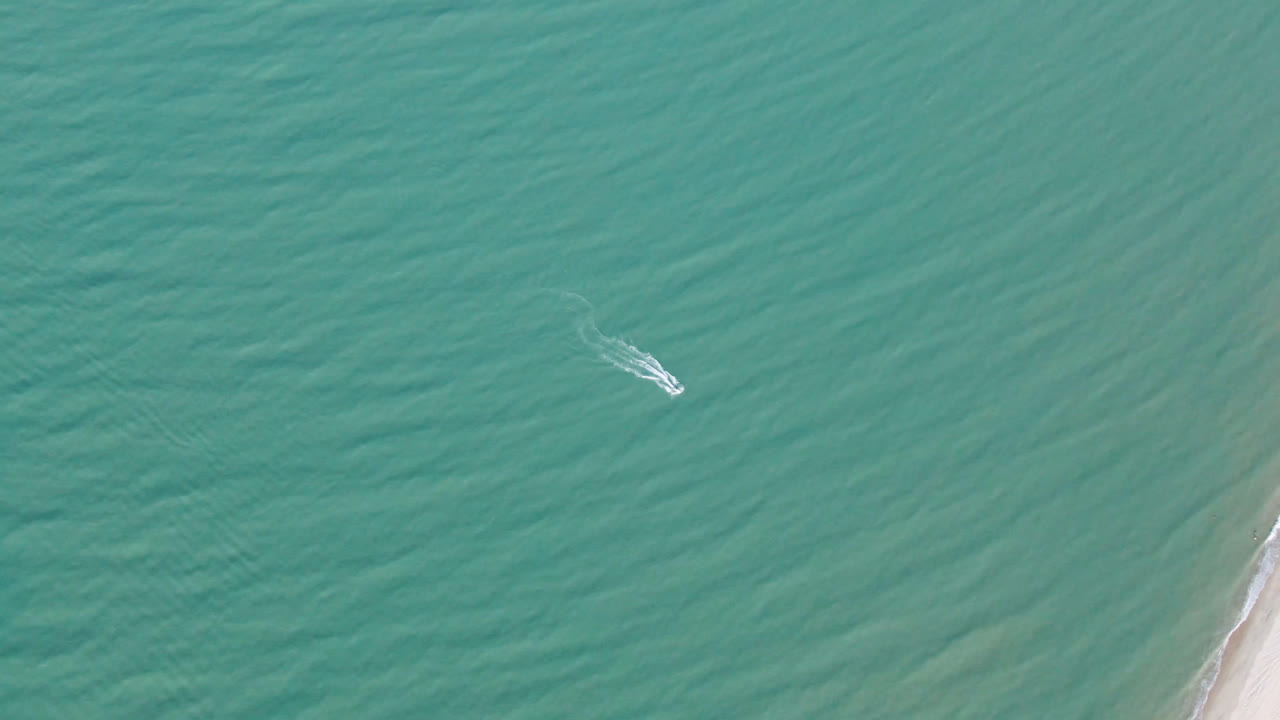 Aerial frame of the small boat slightly farther across green-blue water, with pale shoreline sand visible at frame right in San Felipe BC, Mexico, and no visible people.