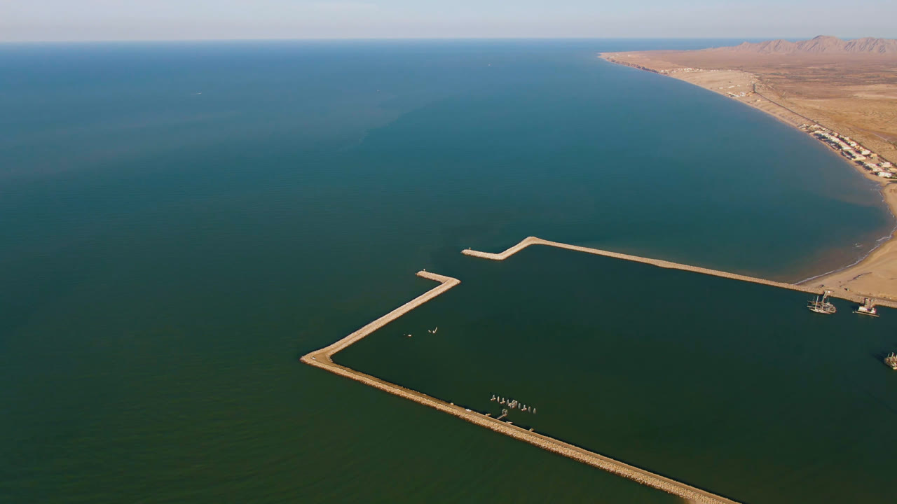 Aerial frame from San Felipe, Baja California, Mexico showing a long angular dock in teal water, sandy coastline to the right, distant mountains, and no visible people.