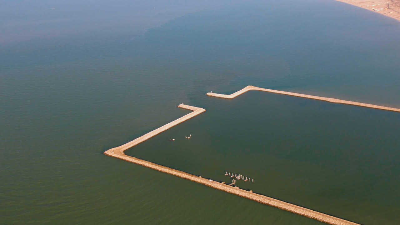 Aerial view in San Felipe with the long dock shifted across the lower frame, small boat shapes near the structure, hazy coast in the background, and no visible people.