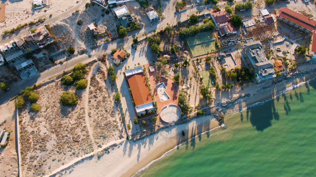 Opening aerial frame over San Felipe BC, Mexico shows a sandy beachfront property with a red-roofed house, tennis court, palm trees, road, and shallow green water, with no visible people.