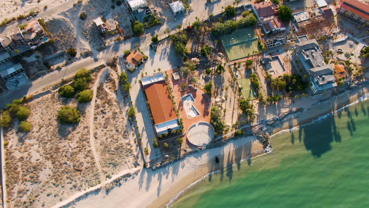 High drone angle over sandy shore, coastal homes, tennis court, and water.