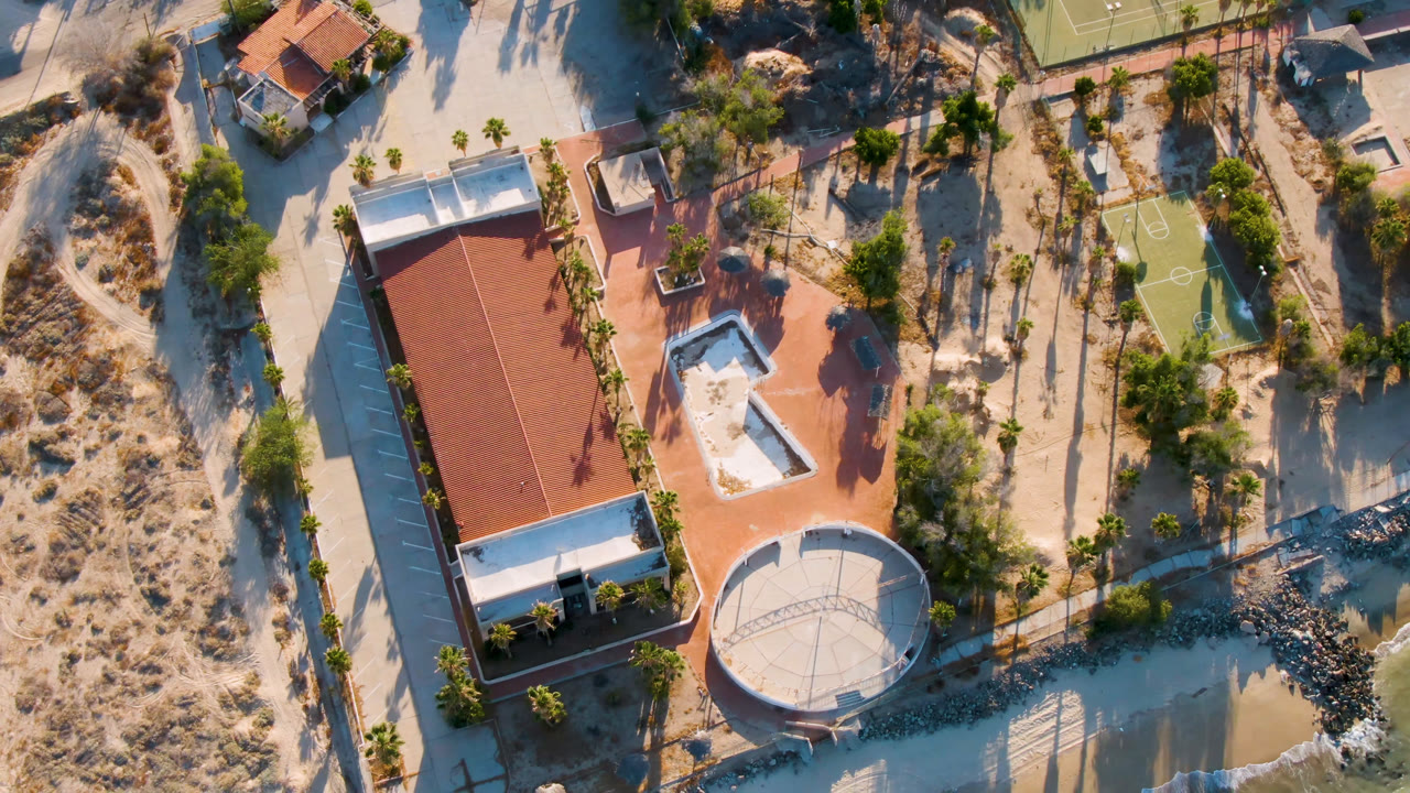 San Felipe overhead frame showing the red-roofed property shifted slightly in view, with the tennis court near the top, sandy paths, water below, and no visible people.