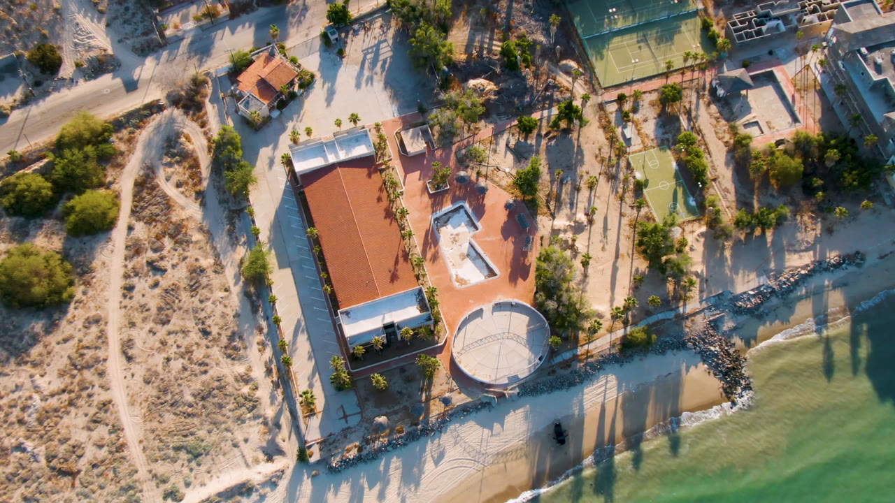 Overhead drone frame in San Felipe, Mexico showing a red-roofed beachside building, round white structure, tennis court, sandy paths, breakwater, green water, and no visible people.