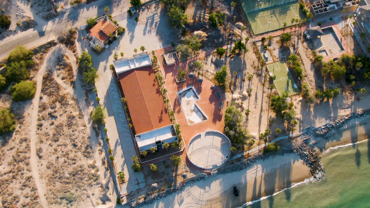 Aerial frame in San Felipe, Mexico with the beachside building centered beside a circular white structure, a tennis court near the top edge, calm water at right, and no visible people.