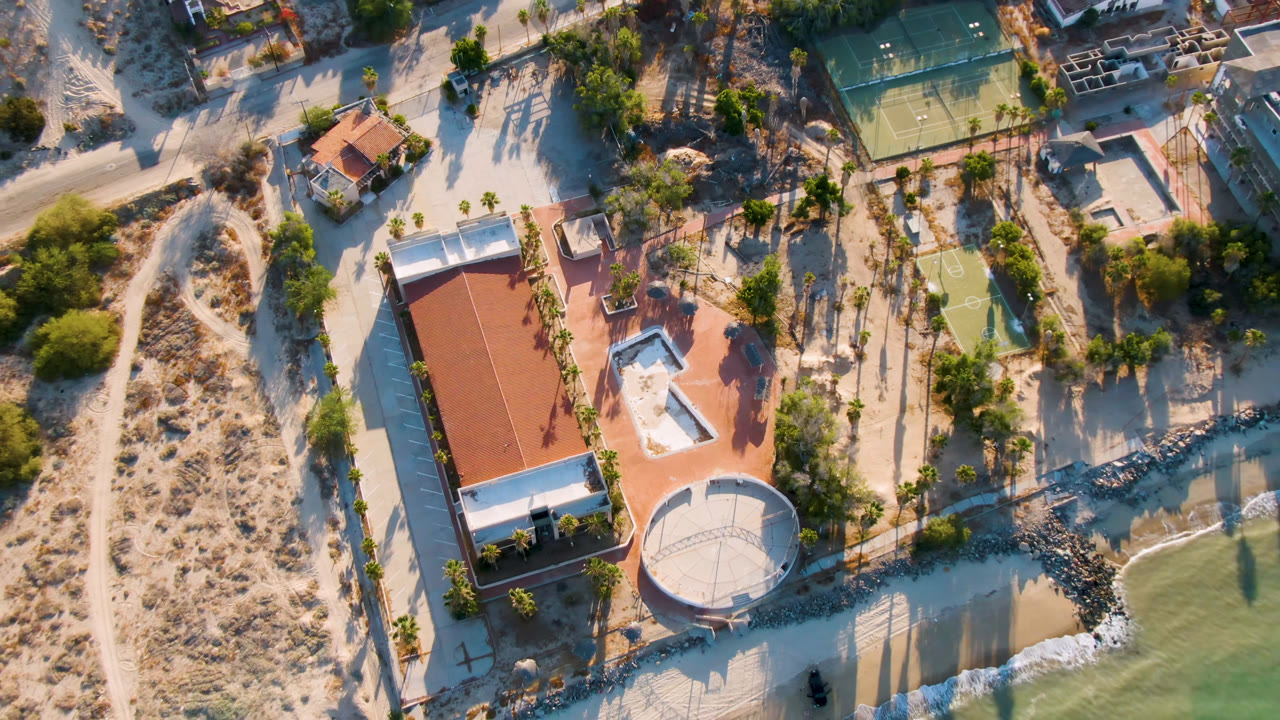 Overhead aerial frame in San Felipe, BC, Mexico showing a red-roofed resort building, round waterfront structure, sandy paths, palm trees, shallow green water, and no visible people.