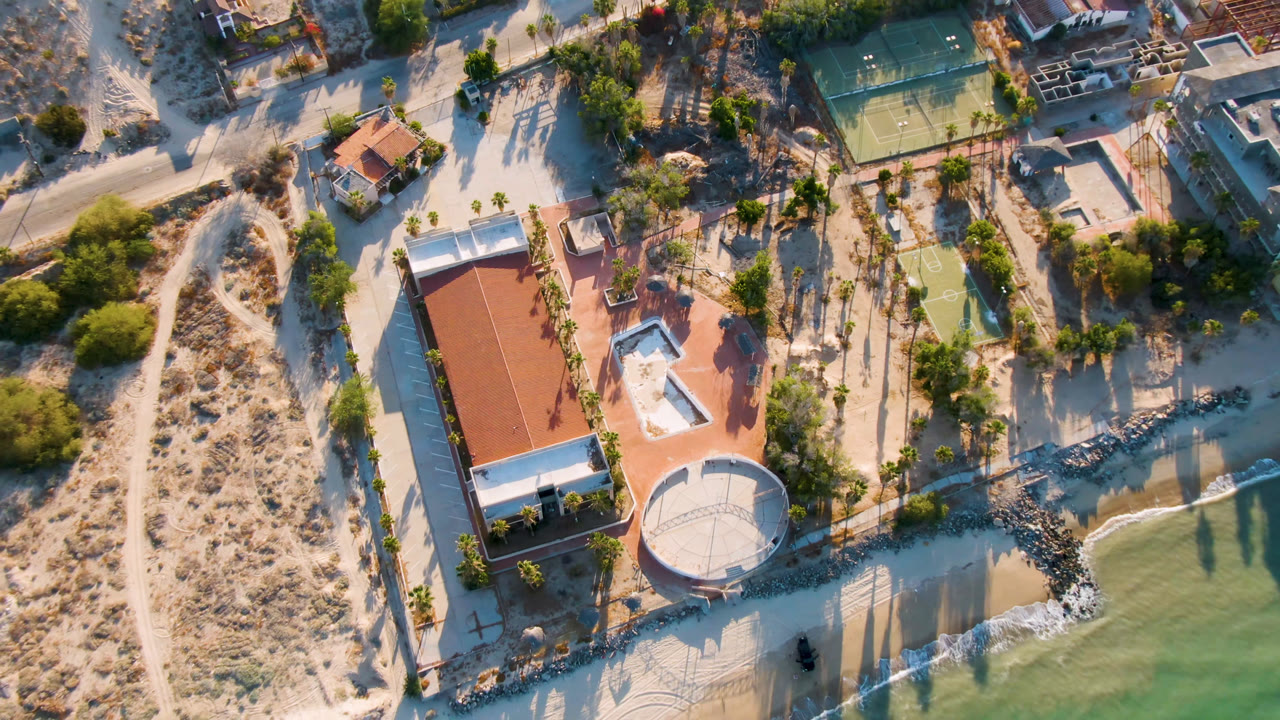 Aerial frame in San Felipe, BC, Mexico with the tennis court near the top, resort roofs at center, palm shadows across sand, shoreline water below, and no visible people.