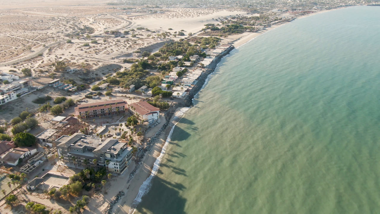 Opening aerial frame over San Felipe, Mexico, showing green-blue water on the right, low shoreline buildings at lower left, dry desert ground inland, and no visible people.