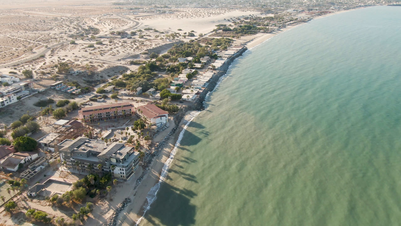 Second aerial frame of San Felipe, Mexico, with the shoreline running diagonally, clustered low buildings near the sand, open green water, dry lots, and no visible people.
