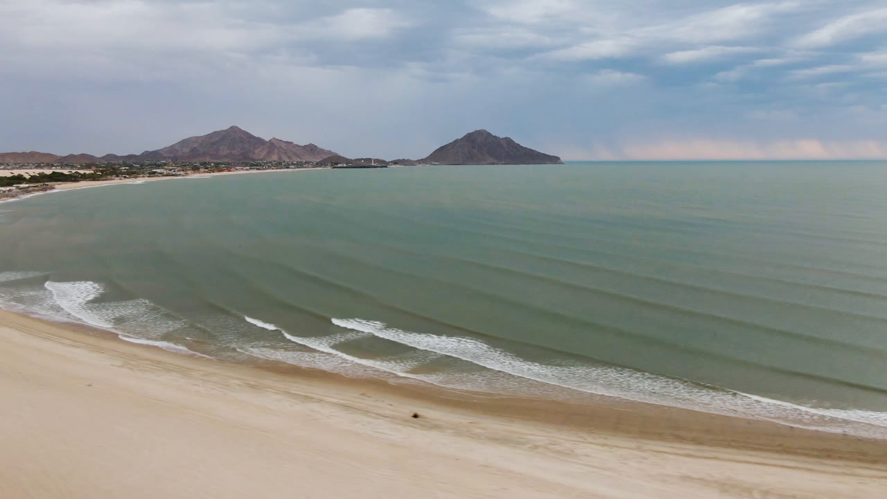 Aerial frame over San Felipe, Baja California, Mexico showing pale sand in the foreground, gentle wave lines across green-blue water, mountains on the horizon, and no visible people.
