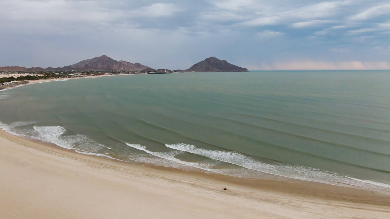 San Felipe aerial view with the shoreline curving from the lower left, thin white surf lines moving toward the sand, distant mountains, cloudy sky, and no visible people.