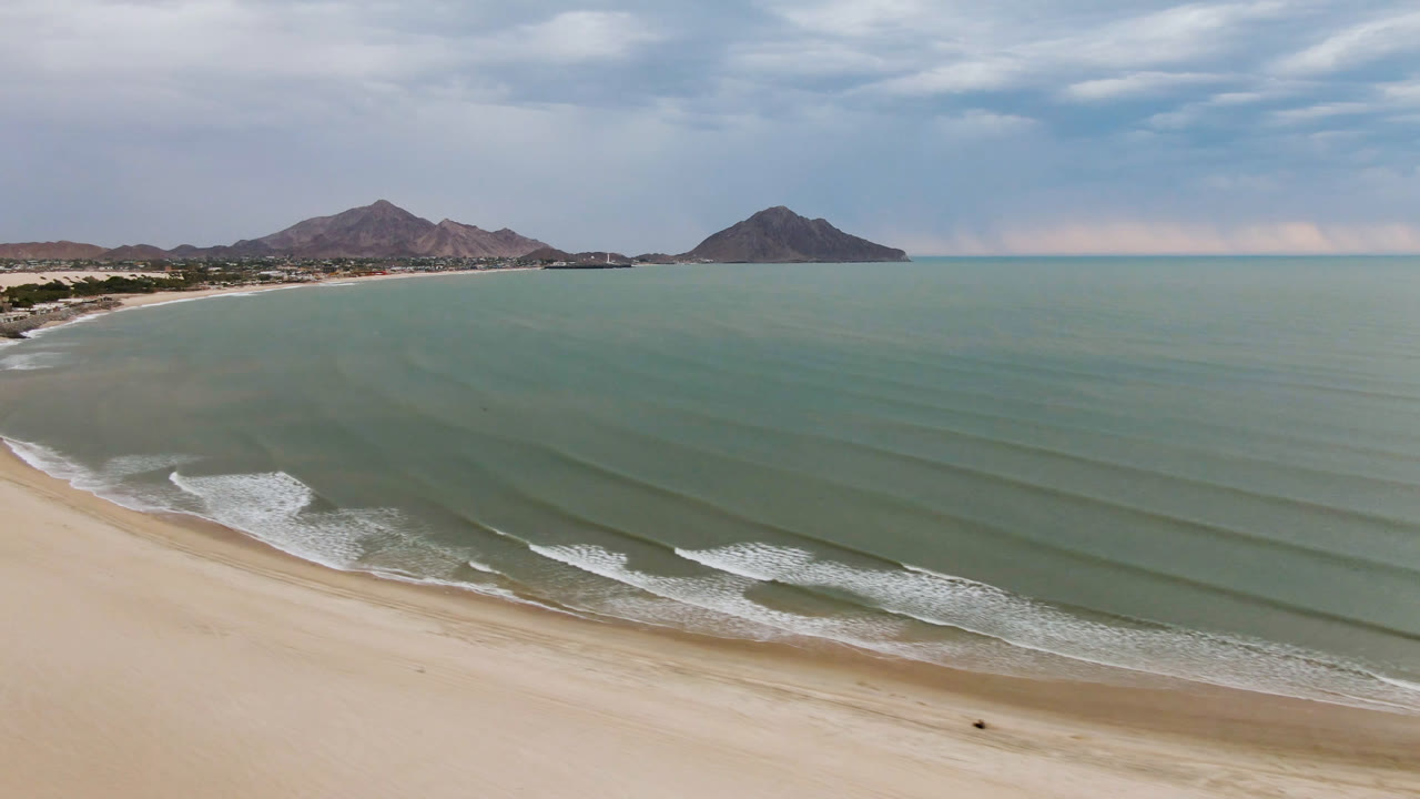 Aerial coastline with empty beach, ocean ripples, mountains, and cloudy sky.