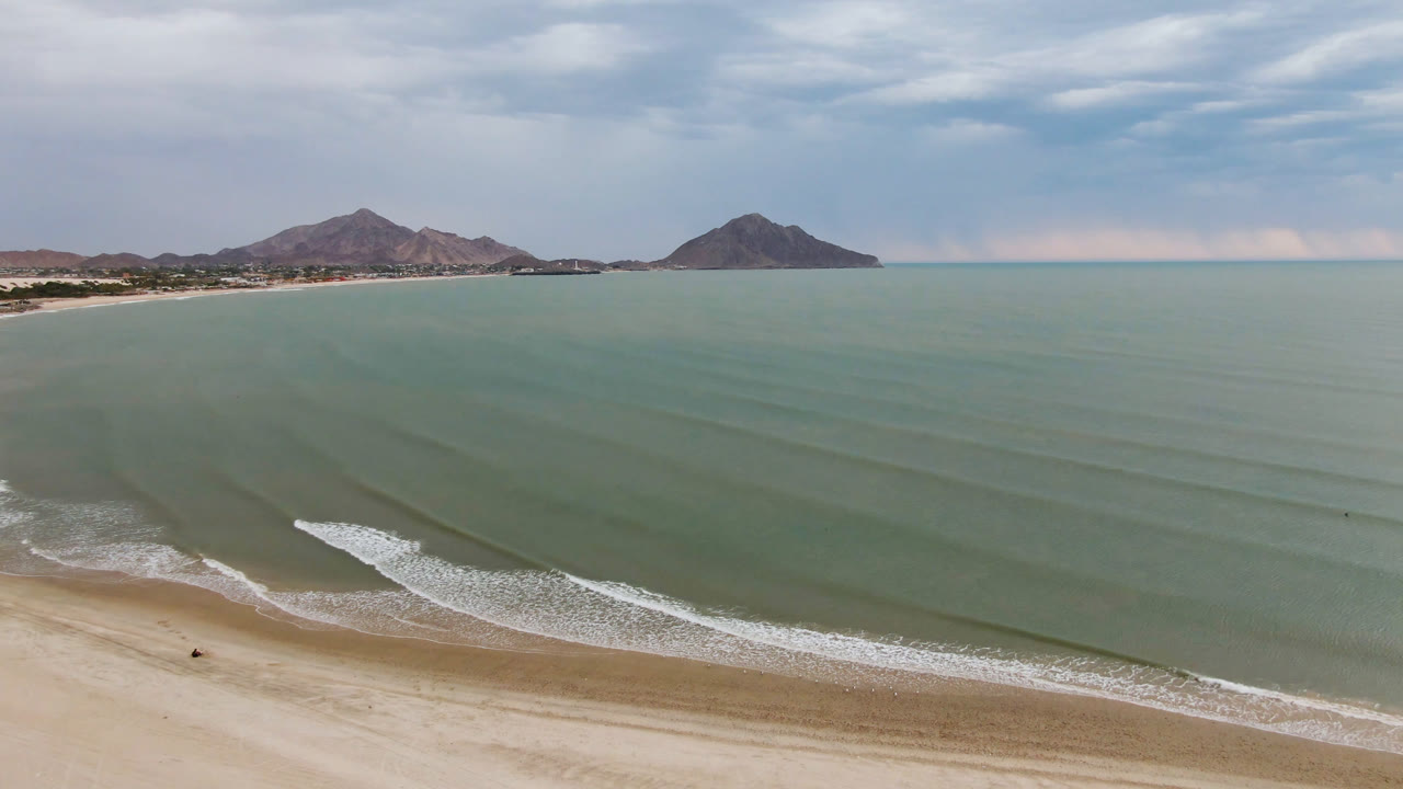 Aerial frame over San Felipe beach in Mexico with pale sand at the bottom, low surf lines across green-blue water, distant mountains on the horizon, and no visible people.