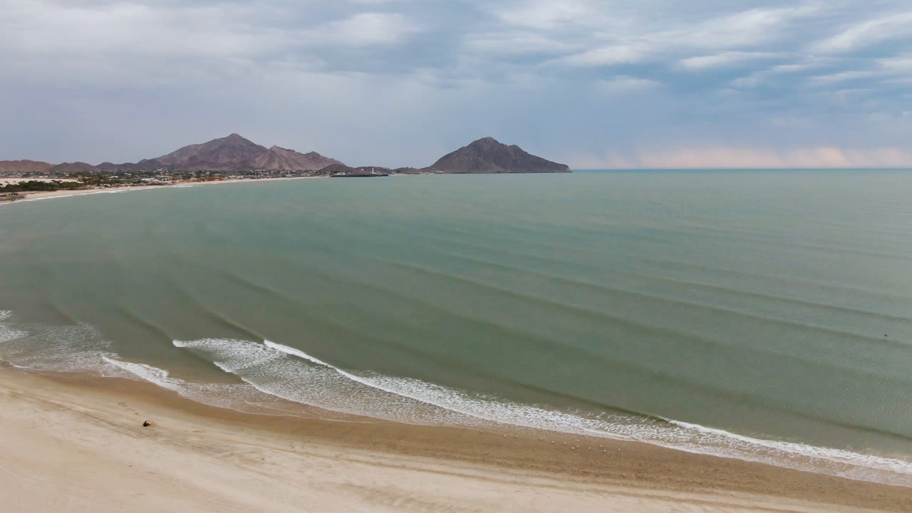 Drone view of a quiet beach with breaking waves and mountain silhouettes.