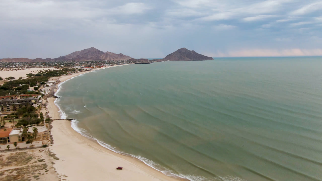 Aerial view of a sandy beach, green ocean water, coastal buildings, and distant mountains.