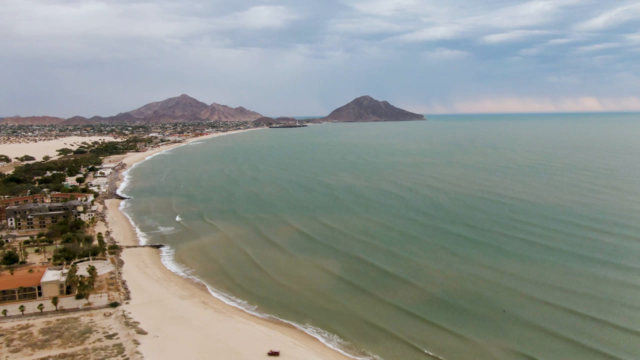 Aerial view in San Felipe, Baja California with the shoreline slightly farther along, small surf lines near the sand, low buildings at lower left, mountains on the horizon, and no visible people.