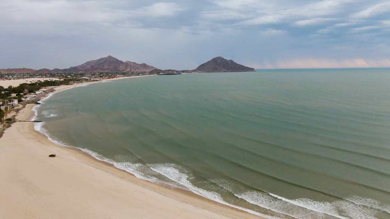 Aerial frame over San Felipe, BC, Mexico, showing more of the curving shoreline at left, shallow green water with parallel wave bands, mountains in the distance, and no visible people.