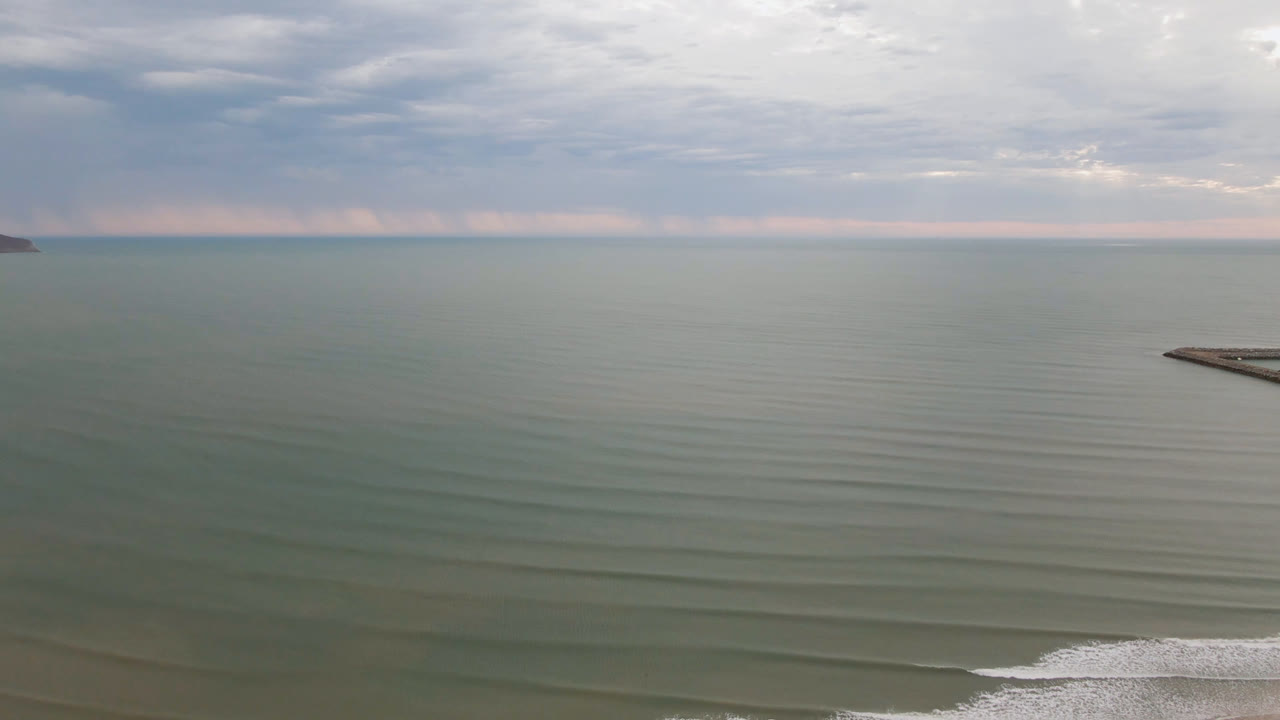 Aerial view in San Felipe with the breakwater more centered to the right, muted green water, layered cloud cover, distant headland, and no visible people.