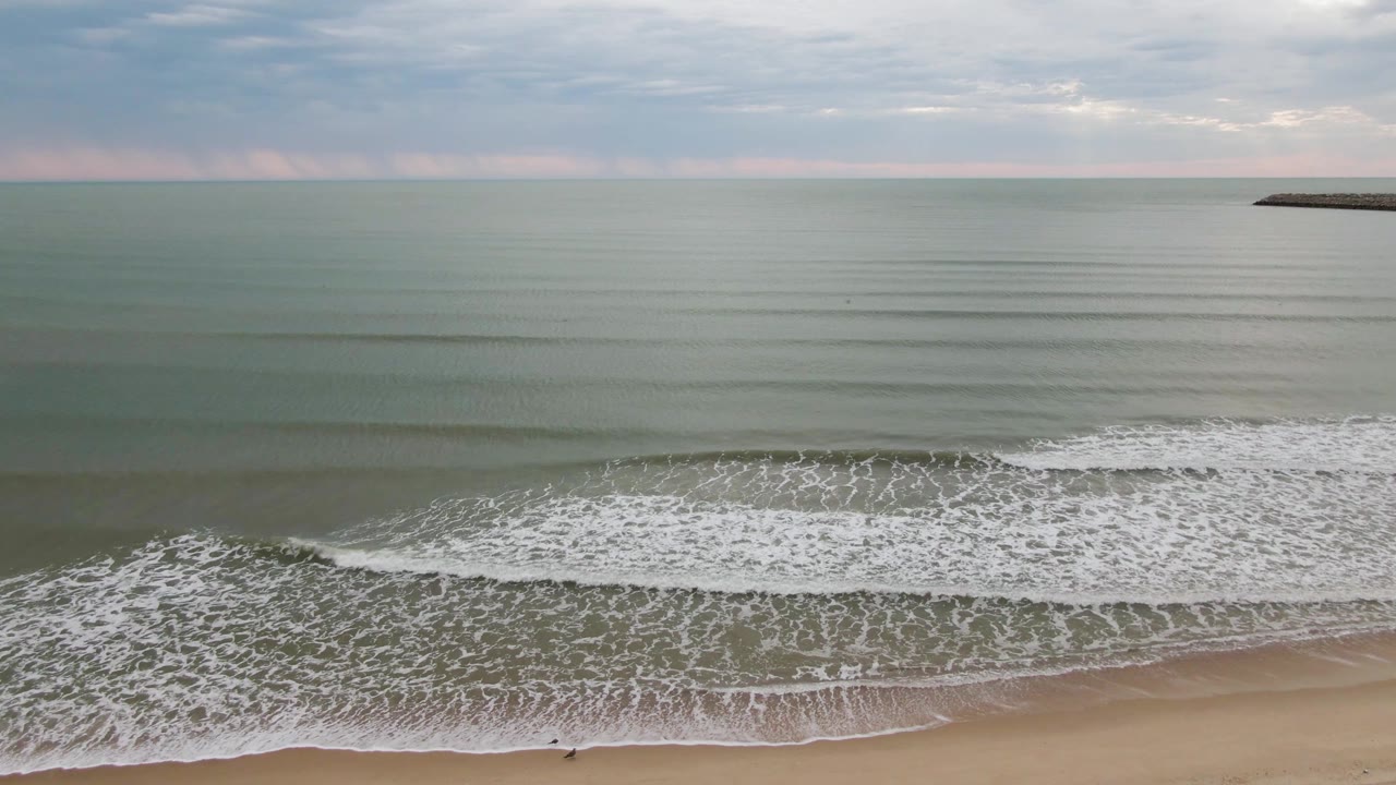Wide San Felipe BC, Mexico beach view with pale sand below, white surf bands across the middle, green-gray ocean to the horizon, and a tiny person-like shape near the shoreline.