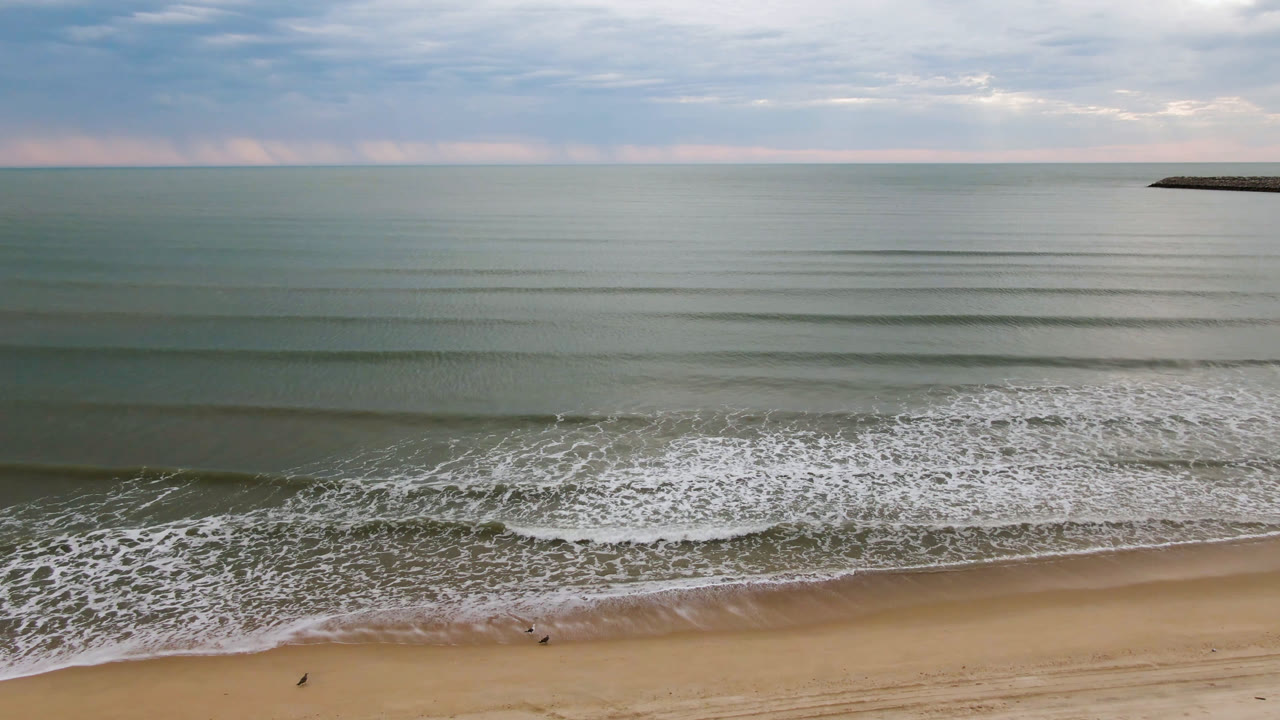 Aerial view of San Felipe, Baja California, Mexico beach with parallel waves reaching tan sand, a cloudy sky above the horizon, and tiny distant beach figures near the shoreline.