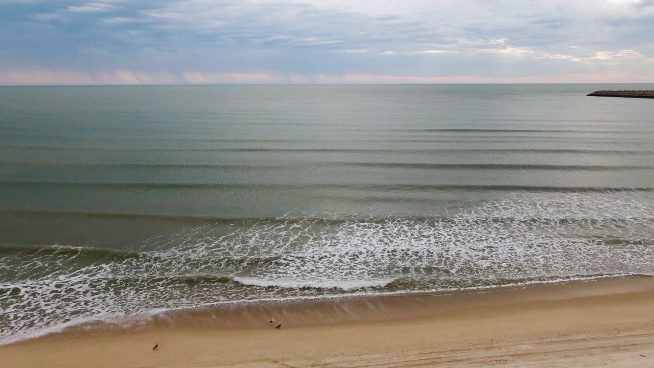 Second aerial moment over San Felipe, Baja California, Mexico shows the surf foam shifted along the sand, with open water, a level horizon, cloudy sky, and tiny distant figures still low in frame.