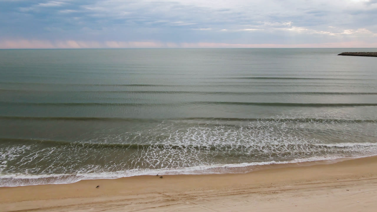 Wide aerial view of San Felipe shoreline at the start of the clip, with pale sand below, small foamy waves across the middle, muted ocean beyond, cloudy sky, and no visible people.
