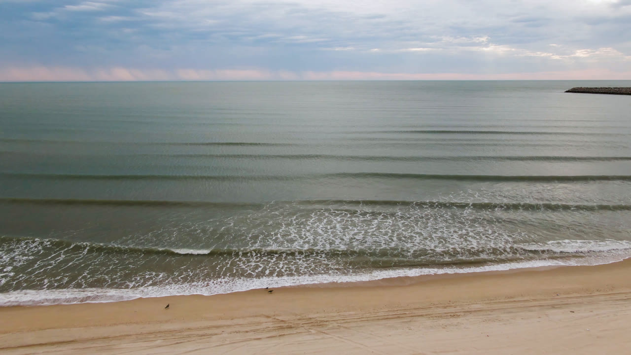 Aerial frame over San Felipe beach one second in, showing parallel bands of gentle surf, open sand at the bottom, calm green-blue water, a distant dark landform near the horizon, and no visible people.