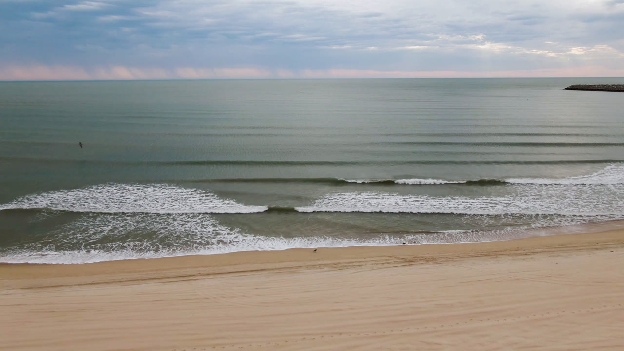 Opening aerial frame of San Felipe, Mexico coastline with tan sand, low surf bands, green-blue water, a cloudy sky, and no visible people.